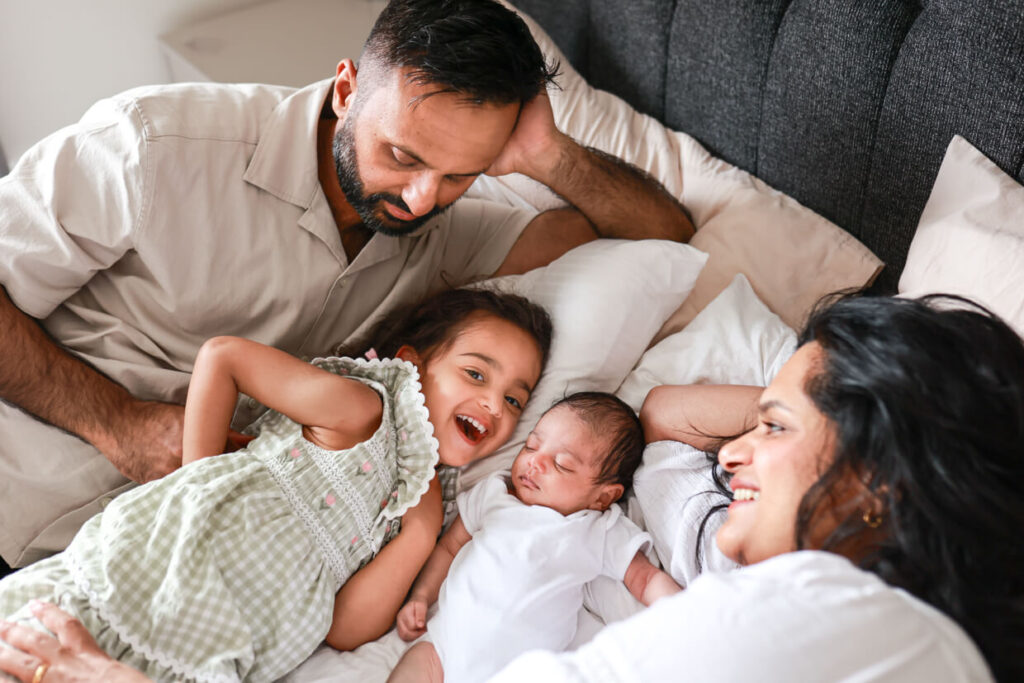 Family of four relaxing together on their bed, with mum leaning against dad as he cuddles their newborn baby