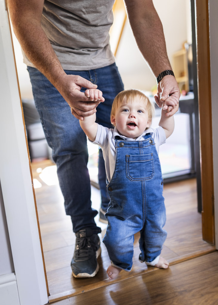 Father holding his toddler's hands as he takes steps through a doorway during an at-home family photoshoot in Glasgow