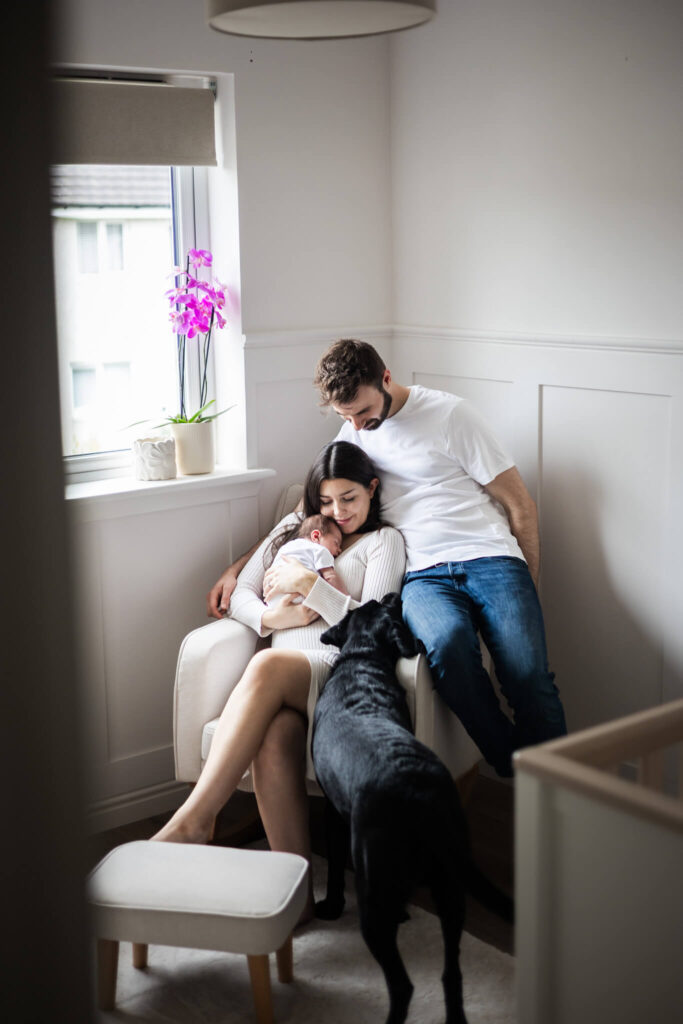 New parents cuddling their newborn baby in a nursing chair with their family dog beside them