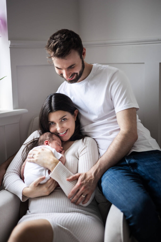 Smiling mum cuddling her newborn in an armchair while dad leans in to hug them both