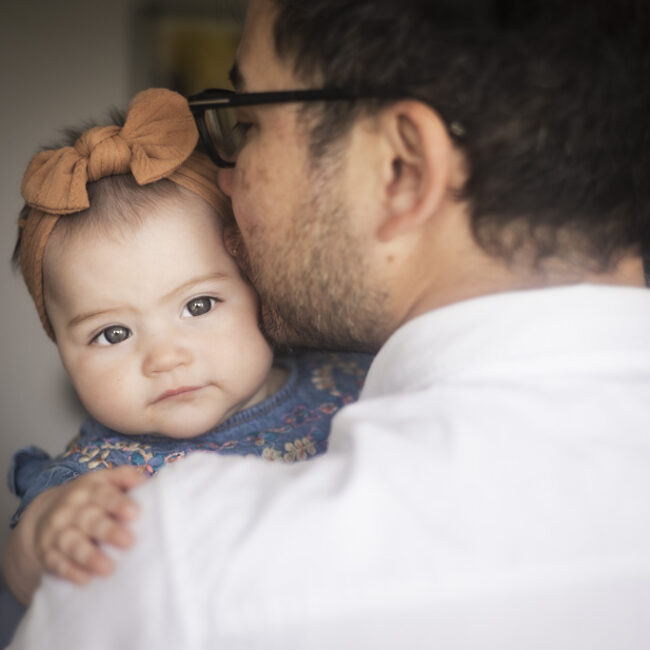 Father wearing glasses kissing his baby daughter's cheek as she looks at the camera during an at-home family photoshoot in Glasgow