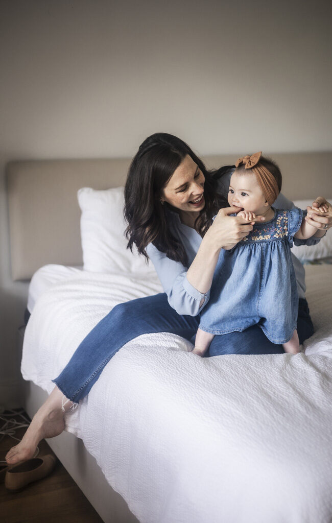 Mother sitting on the bed and playfully holding her smiling baby daughter in a denim dress during an at-home family photoshoot in Glasgow